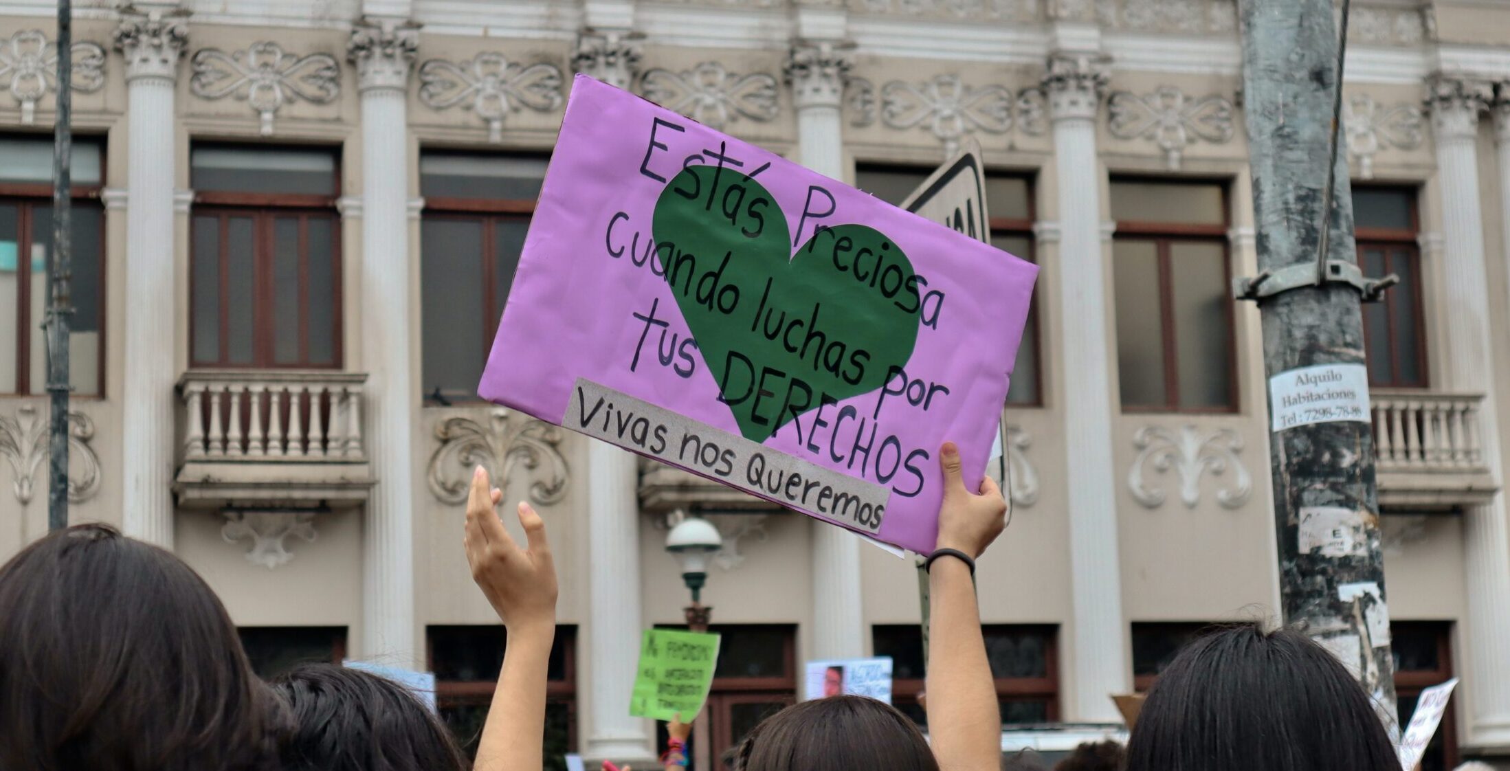 Image of a park with a woman holding a sign, states "Estás Preciosa. Vivas nos queremos"